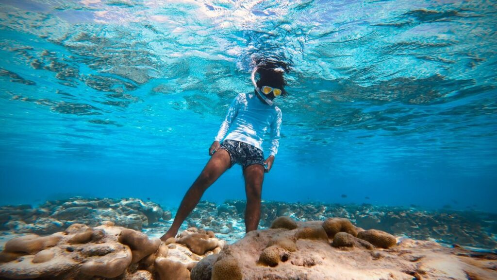 From above full body of anonymous person In snorkeling mask wearing white cardigan and blue shorts standing underwater and examining bottom of transparent sea