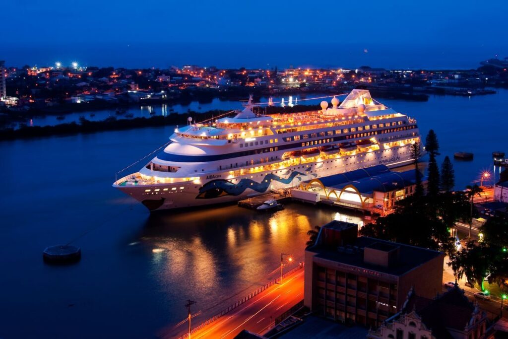 Guía de cruceros por el Caribe en 2026 A brightly lit cruise ship docked at night, reflecting on calm river waters, surrounded by city lights.